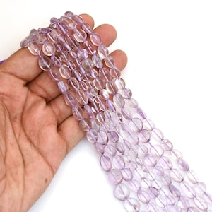 Può includere: Un filo di perline di ametista ovali, traslucide e di colore viola chiaro. Le perline sono lisce, levigate e infilate in una linea continua, mostrando una tonalità viola pallido costante e una consistenza superficiale riflettente e vitrea.