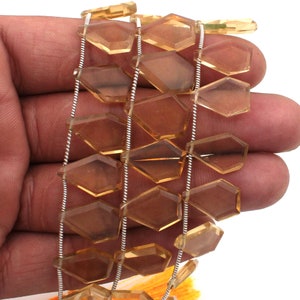 Può includere: Una fila di gemme di citrino sfaccettate a forma di aquilone con una tonalità giallo oro traslucida. Le pietre sono disposte in un motivo verticale, infilate su un sottile filo color argento, mettendo in risalto il taglio geometrico.