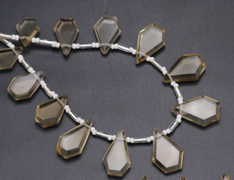Puede incluir: Una hebra de cuentas de piedras preciosas de color marr&oacute;n claro con forma de l&aacute;grima facetada. Las cuentas est&aacute;n enhebradas en una cadena plateada con cuentas espaciadoras blancas.