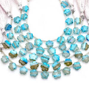 Peut inclure: Quatre rangs de perles en forme de fleur bleu clair et brun translucide disposés en arc. Chaque perle présente une surface à facettes et est séparée par de fines entretoises en métal argenté. Des pompons blancs sont fixés aux extrémités.