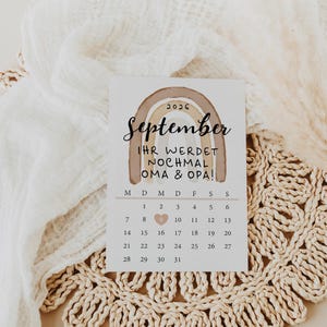 May include: A calendar card for September 2026 with a brown rainbow design. The text reads "IHR WERDET NOCHMAL OMA & OPA!" with a heart marking the 9th. The card is on a woven placemat.