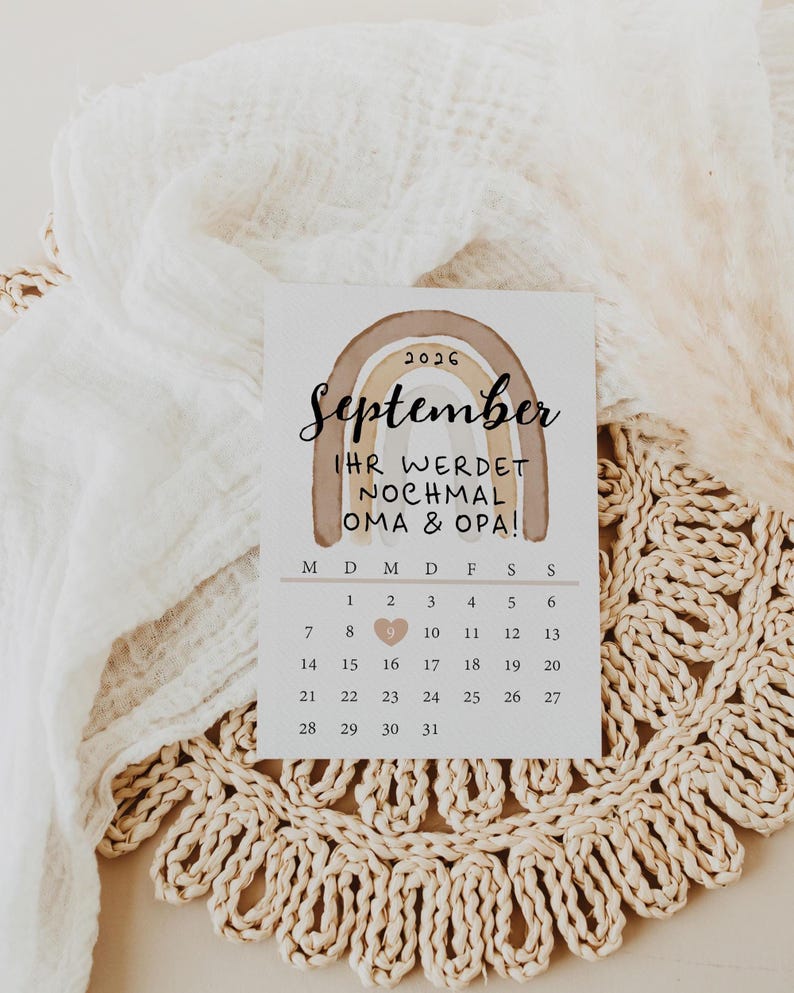 May include: A calendar card for September 2026 with a brown rainbow design. The text reads "IHR WERDET NOCHMAL OMA & OPA!" with a heart marking the 9th. The card is on a woven placemat.