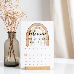 Eine minimalistische Karte zur Schwangerschaftsverkündigung mit einem Kalender für Februar 2026, einem braunen Regenbogen im Aquarellstil und der Aufschrift „Wir sind bald zu dritt”. Die Karte steht auf einem Tisch und wird in einen Holzständer gelegt. Im Hintergrund steht eine Vase mit getrockneten Blumen, und eine Hängegardine hängt herunter.