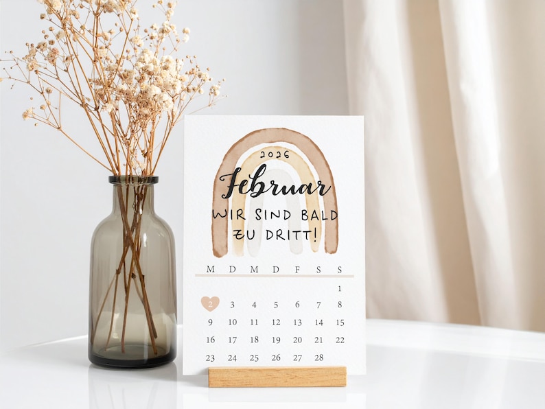 Eine minimalistische Karte zur Schwangerschaftsverkündigung mit einem Kalender für Februar 2026, einem braunen Regenbogen im Aquarellstil und der Aufschrift „Wir sind bald zu dritt”. Die Karte steht auf einem Tisch und wird in einen Holzständer gelegt. Im Hintergrund steht eine Vase mit getrockneten Blumen, und eine Hängegardine hängt herunter.
