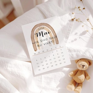 May include: A May 2026 calendar with a watercolor rainbow design and the text "WIR SIND BALD ZU VIERT!" sits on a white surface. A small teddy bear rests nearby. The calendar is a light beige color.