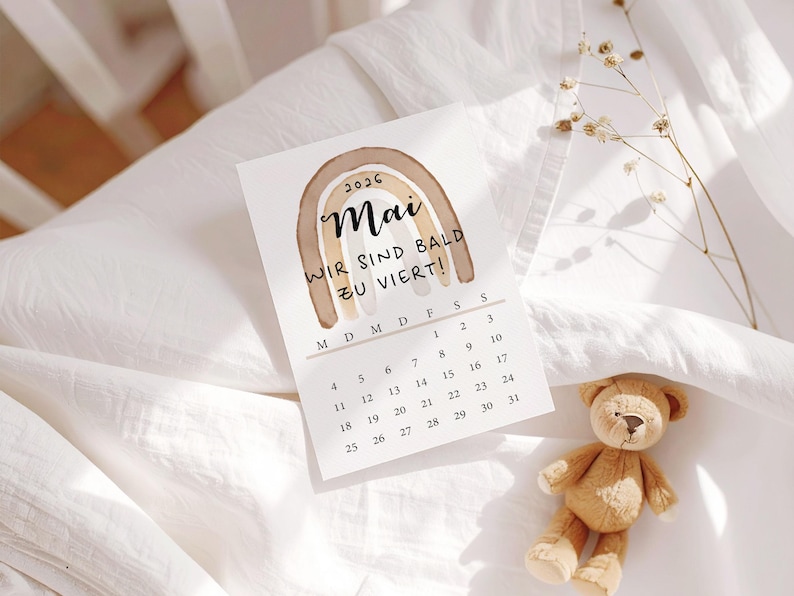 May include: A May 2026 calendar with a watercolor rainbow design and the text "WIR SIND BALD ZU VIERT!" sits on a white surface. A small teddy bear rests nearby. The calendar is a light beige color.