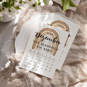 May include: Two announcement cards with a watercolor rainbow design and the text "Dezember WIR BEKOMMEN EIN BABY!" are displayed on a white plate. The cards have a calendar for December 2026. The background includes a beige fabric and white flowers.