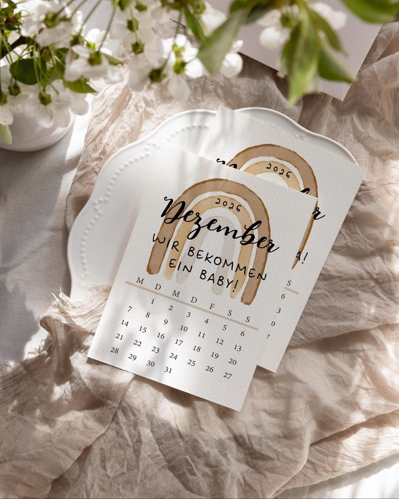 May include: Two announcement cards with a watercolor rainbow design and the text "Dezember WIR BEKOMMEN EIN BABY!" are displayed on a white plate. The cards have a calendar for December 2026. The background includes a beige fabric and white flowers.