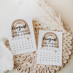 May include: Two August 2026 announcement cards with a watercolor rainbow design. The cards have a calendar layout and text in German, announcing the arrival of a baby. The cards are on a cream-colored, crocheted surface.