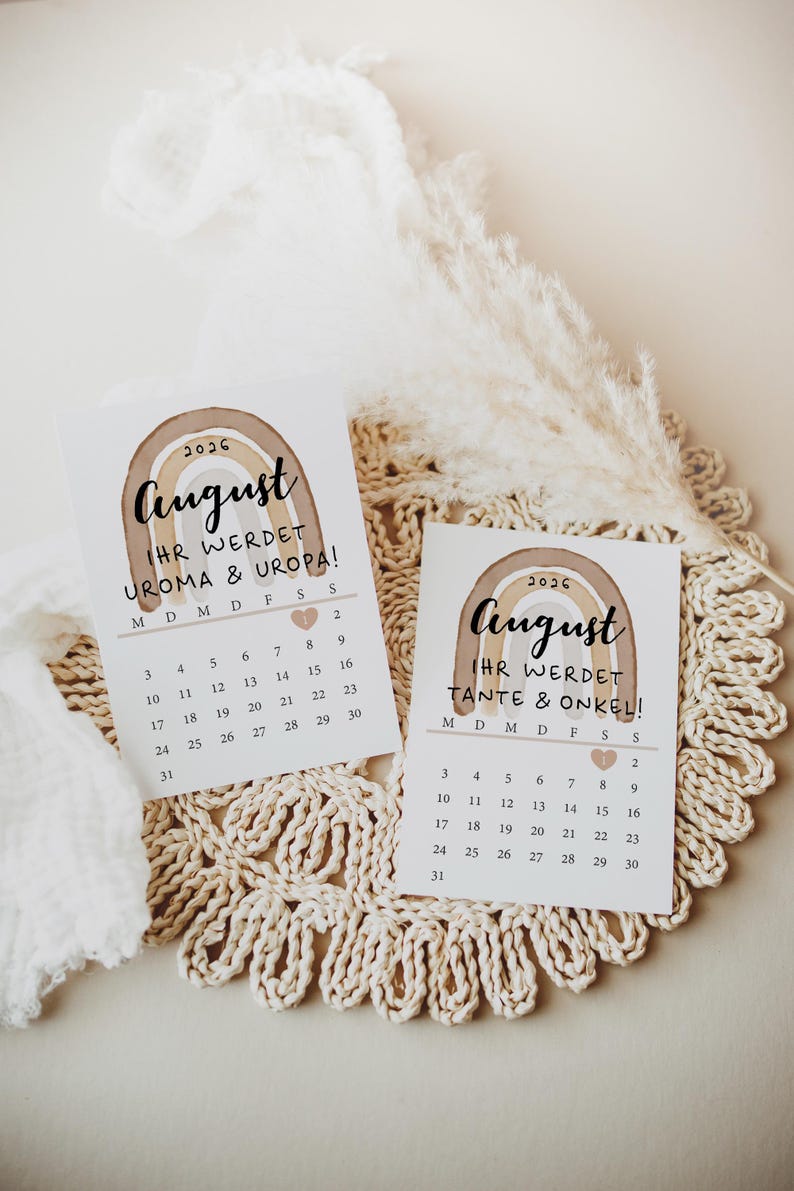 May include: Two August 2026 announcement cards with a watercolor rainbow design. The cards have a calendar layout and text in German, announcing the arrival of a baby. The cards are on a cream-colored, crocheted surface.