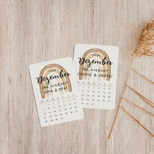 May include: Two small, rectangular cards with a watercolor rainbow design and the word "Dezember" in script. The cards display a calendar for December 2026, with the text "IHR WERDET OMA & OPA!" and "UROMA & UROPA!" in German.