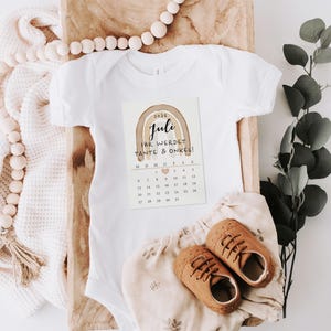 May include: A white baby onesie with a printed card featuring a watercolor rainbow and the text "Juli Ihr werdet Tante & Onkel!". Brown leather baby shoes and a floral print diaper cover are also present.