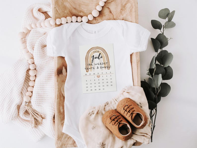 May include: A white baby onesie with a printed card featuring a watercolor rainbow and the text "Juli Ihr werdet Tante & Onkel!". Brown leather baby shoes and a floral print diaper cover are also present.