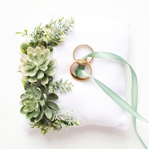 Peut inclure: Un oreiller blanc avec un ruban vert et deux alliances dor&eacute;es sur le dessus. L'oreiller est d&eacute;cor&eacute; de succulentes vertes et de verdure.