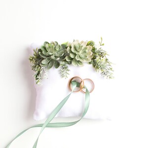 Peut inclure: Oreiller blanc avec une composition florale de succulentes vertes et deux alliances dor&eacute;es sur le dessus. Un ruban vert sauge est nou&eacute; autour de l'oreiller.