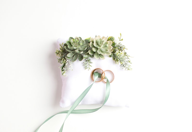 Peut inclure: Oreiller blanc avec une composition florale de succulentes vertes et deux alliances dor&eacute;es sur le dessus. Un ruban vert sauge est nou&eacute; autour de l'oreiller.