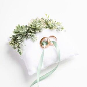 Peut inclure: Oreiller blanc avec des accents floraux de succulentes vertes et deux alliances dor&eacute;es attach&eacute;es avec un ruban vert sauge.