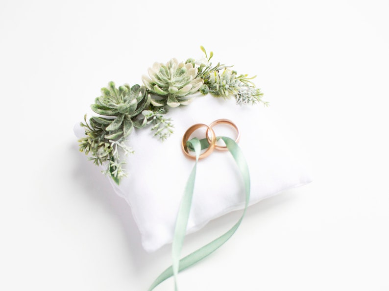Peut inclure: Oreiller blanc avec des accents floraux de succulentes vertes et deux alliances dor&eacute;es attach&eacute;es avec un ruban vert sauge.