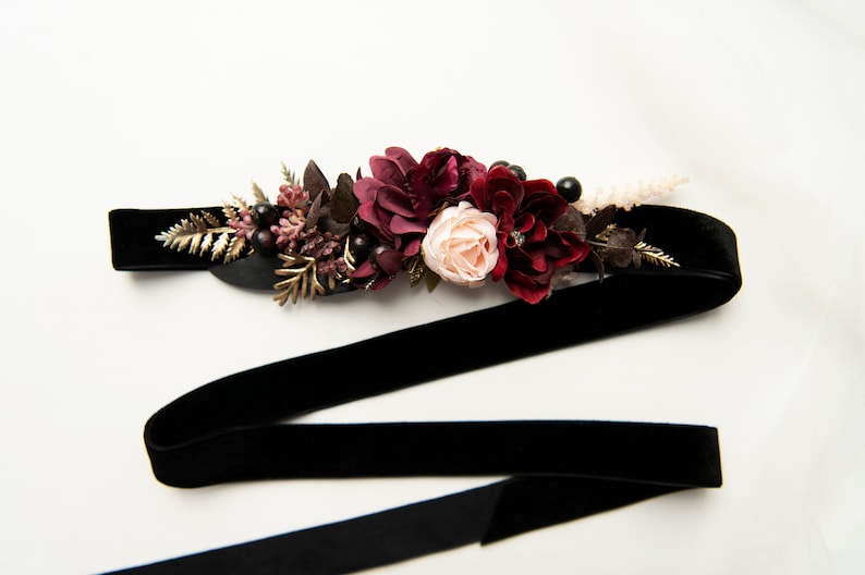 May include: A black velvet ribbon with a floral embellishment. The flowers are burgundy, pink, and white. The ribbon is tied in a bow.