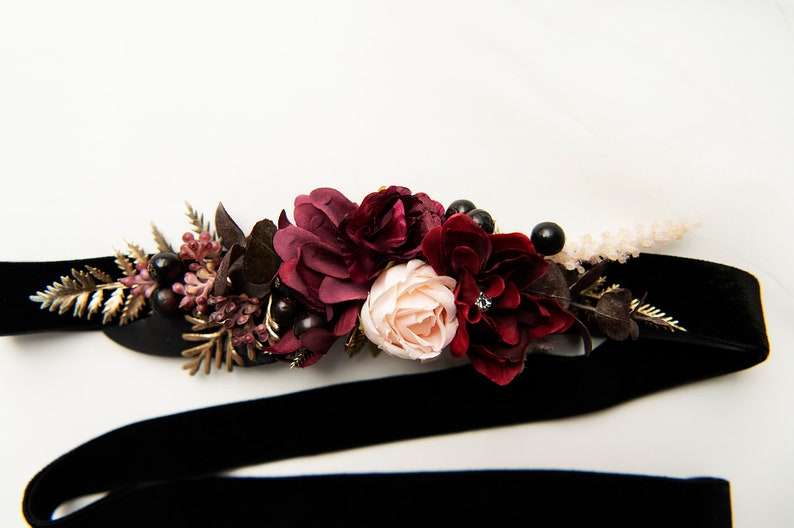 May include: A black velvet ribbon with a burgundy and blush floral arrangement. The arrangement includes burgundy roses, blush roses, burgundy berries, and gold leaves.