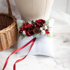 May include: White ring pillow with a red and white floral arrangement. The arrangement includes red roses, berries, pine cones, and white beads. The pillow has a red satin ribbon.