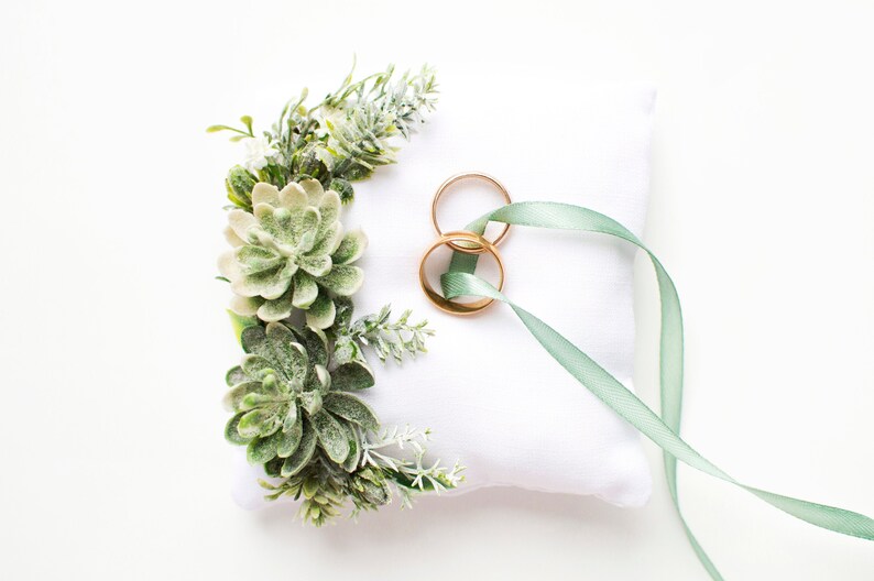 Peut inclure: Un oreiller blanc avec un ruban vert et deux alliances dor&eacute;es sur le dessus. L'oreiller est d&eacute;cor&eacute; de succulentes vertes et de verdure.