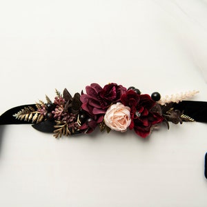 May include: A black velvet ribbon with a burgundy and blush floral embellishment. The floral arrangement includes burgundy roses, blush roses, and burgundy berries.
