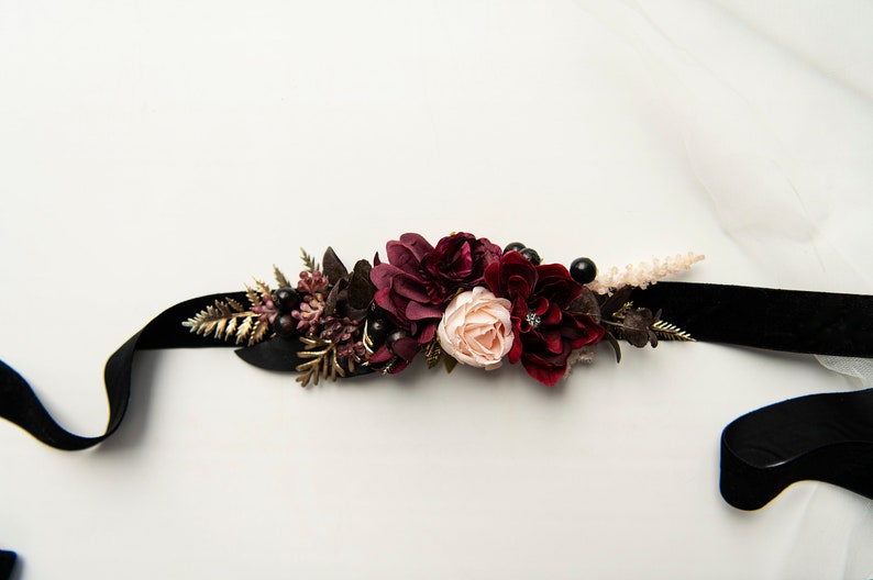 May include: A black velvet ribbon with a burgundy and blush floral embellishment. The floral arrangement includes burgundy roses, blush roses, and burgundy berries.