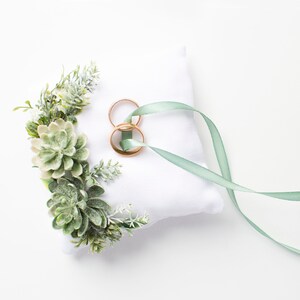 Peut inclure: Un oreiller blanc avec une composition florale de succulentes vertes et un ruban vert nou&eacute; autour de deux alliances dor&eacute;es.