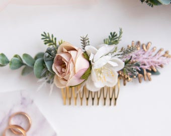 May include: A floral hair comb featuring a blush rose, white flowers, and greenery. The comb has gold-colored teeth and is adorned with sprigs of lavender and gold accents. Wedding hair accessory.