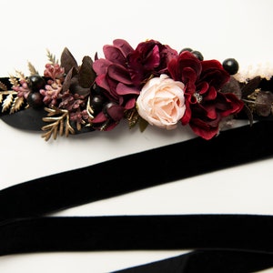 May include: A black velvet ribbon with a burgundy and blush floral embellishment. The floral arrangement includes burgundy roses, blush roses, and burgundy berries.