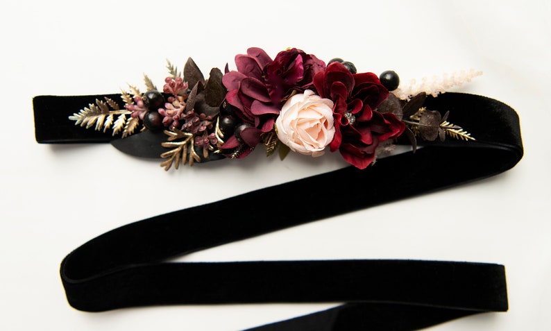 May include: A black velvet ribbon with a burgundy and blush floral embellishment. The floral arrangement includes burgundy roses, blush roses, and burgundy berries.