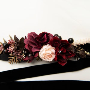 May include: A black velvet ribbon with a burgundy and blush floral arrangement. The arrangement includes burgundy roses, blush roses, burgundy berries, and gold leaves.