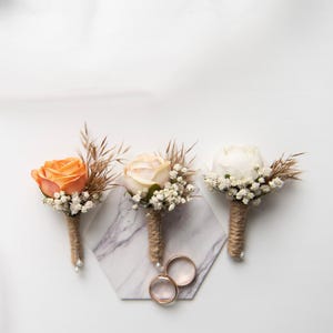 Puede incluir: Tres boutonnières con rosas de diferentes colores y aliento de bebé blanco sobre una superficie de mármol con dos anillos de oro.