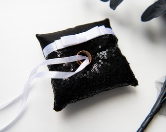 May include: A black sequined pillow with white satin ribbon and bow, with two gold wedding bands on top.