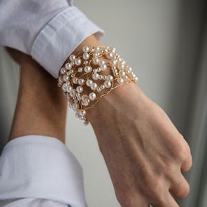 May include: A gold wire bracelet with white pearl beads. The bracelet is wrapped around a wrist.