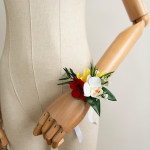 Op de afbeelding: Een bloemencorsage voor om de pols met een witte orchidee, een donkerrode bloem en gele bloemblaadjes, geaccentueerd met groene bladeren en kleine witte vulbloemen. Het geheel is bevestigd met een wit lint om de pols van een houten etalagepop.