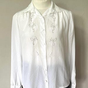 May include: A white blouse with a collared neckline and long sleeves. It has an embroidered design down the front and on the shoulders. The fabric has a subtle dotted texture. The blouse is buttoned up.