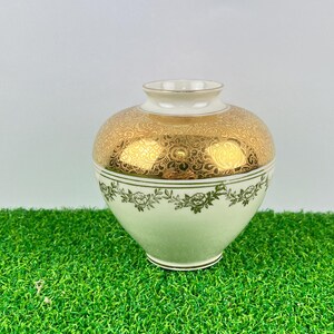 May include: A decorative vase with a bulbous shape. The top half is gold with an ornate pattern, while the bottom half is a pale green with a floral design. The rim and the top of the vase are white.