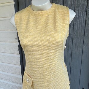 May include: A sleeveless, yellow dress with a textured pattern. The dress has a round neckline, a small pocket, and buttons down the side. The dress is displayed on a mannequin.