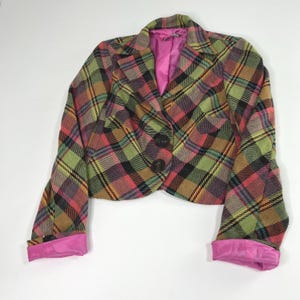 May include: A cropped blazer featuring a multi-coloured plaid pattern in shades of green, pink, tan, and black. The jacket has a notched lapel, two dark brown buttons on the front, and long sleeves with bright pink lining visible at the cuffs.