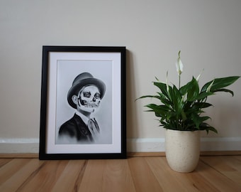 Könnte beinhalten: Ein gerahmtes Schwarz-Weiß-Porträt einer Person mit Totenkopf-Gesichtsbemalung, die einen Hut, einen Anzug und eine Krawatte trägt. Das Kunstwerk wird auf einem Holzboden neben einer Topfpflanze mit Spathiphyllum in einem gesprenkelten weißen Topf ausgestellt.