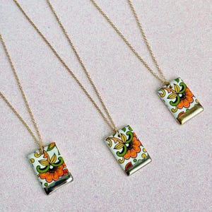 May include: Three gold-tone necklaces with rectangular pendants. Each pendant features a floral design in orange, yellow, and green on a white background. The pendants have a gold-colored metal border. The necklaces are displayed on a pink surface.