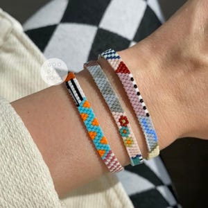 May include: Three colorful beaded bracelets with geometric patterns in shades of blue, orange, pink, white, and gray. The bracelets are stacked on a person's wrist.
