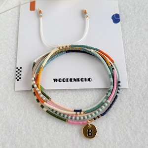 May include: A set of colourful beaded bracelets. The bracelets are made of small beads in shades of pink, green, blue, and orange. A gold charm with the letter "B" is attached to one. The brand name "WOODENBOHO" is printed on the card.