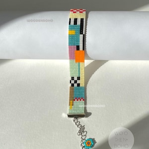 May include: A colourful beaded bracelet with a geometric pattern in shades of blue, green, yellow, orange, pink, black and white. The bracelet has a silver clasp and a small blue and orange flower charm.
