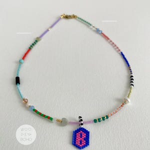 Puede incluir: Un collar de cuentas de colores con un colgante de cuentas azul y rosa en forma de ocho. El collar está hecho con una variedad de cuentas, incluyendo cuentas rojas, naranjas, amarillas, verdes, azules, moradas y blancas. El colgante está unido al collar con un cierre dorado.