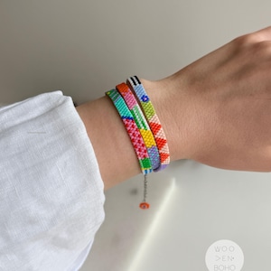 以下が含まれることがあります： 手首に3つのカラフルなビーズブレスレット。ブレスレットは、ピンク、緑、青、黄色など、さまざまな模様と色を特徴としています。ブレスレットは、小さなオレンジ色のビーズが付いたシルバーチェーンで接続されています。