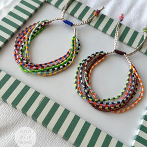 Può includere: Due braccialetti a più fili realizzati con piccole perline colorate. Ogni braccialetto presenta diversi fili di perline in varie tonalità, tra cui verde, blu, arancione e giallo, fissati con cordini beige intrecciati e nodi scorrevoli regolabili.
