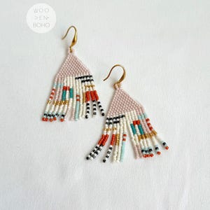 Può includere: Un paio di orecchini a forma di frange in perle rosa con un motivo geometrico in bianco, nero, rosso, turchese e oro. Gli orecchini sono realizzati con piccole perle.